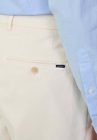 Pantaloni di cotone beige chiaro con una tasca posteriore e chiusura a bottone singolo, abbinati a una camicia a quadri azzurri. Etichetta del brand visibile.