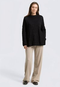 Schwarzer Strickpullover mit langen Ärmeln, hohem Kragen und Seitentaschen, kombiniert mit beigen weit geschnittenen Hosen und schwarzen Schuhen.