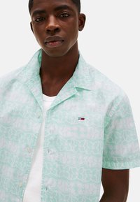 Camisa de manga corta color verde menta con patrón abstracto blanco, cierre de botones y pequeña etiqueta de logo en el pecho. Tejido de algodón texturizado.