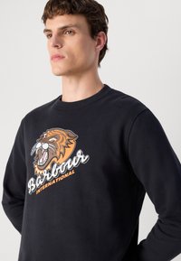 Fekete pulóver hosszú ujjal, melyen egy feltűnő narancssárga és szürke oroszlán grafika és fehér szöveg látható, amely így olvasható: "Barbour International."