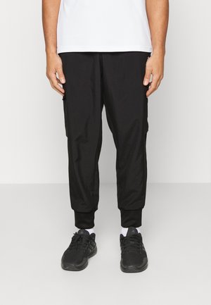 Pantalons noirs fuselés en tissu léger, avec des poignets élastiques et des poches latérales, associés à des chaussures de sport noires et des chaussettes blanches.
