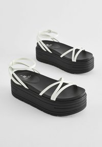 Zwarte platform sandalen met witte bandjes, voorzien van een gladde bovenkant en een gestructureerde zool. Inclusief verstelbare enkelbandjes.