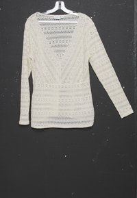 Blouse en dentelle blanche à manches longues avec col en V, suspendue sur un cintre blanc contre un fond texturé sombre.