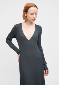 Φόρεμα με μακριά μανίκια και ριμπ υφή σε σκούρο γκρι. Βαθύ V ντεκολτέ, εφαρμοστό κορσέ και ιριδίζουσα φούστα. Μαλακή υφή με διακριτικό μοτίβο.