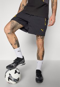 Sorte atletiske shorts med gule accenter, båret med sorte fodboldstøvler. Fodbold på jorden, med et sort og hvidt design.