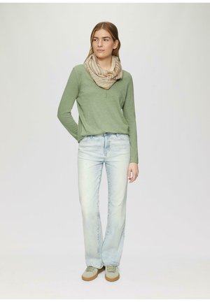 Jeune femme debout portant un haut vert à manches longues, une écharpe beige rayée, un jean bleu clair et des baskets vertes sur un fond uni.