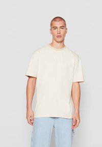 Beige cotton t-shirt with a crew neckline and short sleeves, featuring subtle white logo embroidery on the chest, paired with light blue jeans.