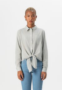 Striped button-up shirt with long sleeves, curved hem, tied at the waist. Worn with light blue jeans, featuring a single chest pocket.