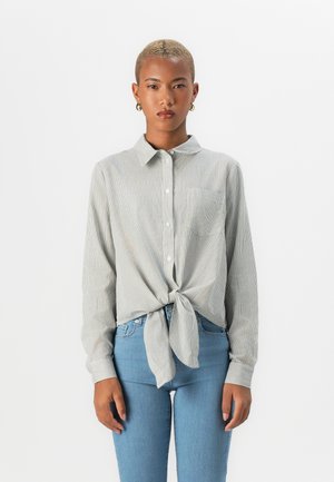 Camicia a righe con maniche lunghe, orlo curvo, legata in vita. Indossata con jeans azzurri chiari, dotati di una tasca sul petto.