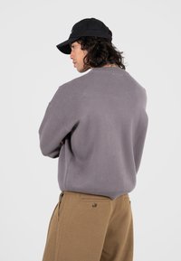 Grauer Strickpullover mit rundem Ausschnitt und lockerer Passform, kombiniert mit beigen Hosen mit Taschen und Knopfdetails. Schwarze Mütze rückwärts getragen.