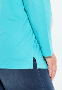 Türkises Langarmhemd mit abgerundetem Saum und seitlichen Schlitzen, aus einem weichen Stoff, präsentiert mit dunklen Jeans.