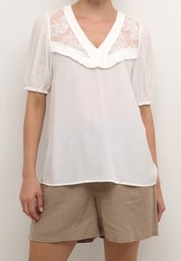 Blusa blanca con yugo bordado transparente, escote en V y ribetes con volantes. Mangas cortas abullonadas y tejido ligero y fluido. Combinada con pantalones cortos beige.