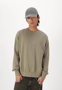 Olijfgroene sweatshirt met lange mouwen, ribbelmanchetten en een ronde hals, gecombineerd met een grijze pet en donkere, gemusterde jeans.