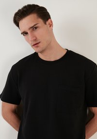 Čierne rebrované tričko s okrúhlym výstrihom a náprsné vrecko. Tkanina má textúrovaný povrch, čo vytvára jemnú vizuálnu hĺbku.