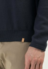 Pull en maille bleu marine avec ourlet et poignets côtelés, doté d'une petite étiquette en cuir marron clair en bas. La texture du tissu est douce et dense.