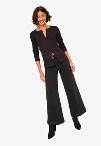 Schwarzer, knöpfbarer Cardigan mit langen Ärmeln und rundem Ausschnitt, kombiniert mit hoch taillierter, weit geschnittener schwarzer Hose und schwarzen Stiefeletten.