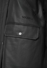 Casaco sintético preto com um bolso com botão, logótipo em relevo e textura suave, destacando um design estruturado.