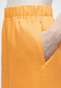 Χέρι στην τσέπη φωτεινών πορτοκαλί παντελονιών με ελαστική μέση και ετικέτα της μάρκας Ecoalf, φορεμένα με λευκό ριμπ τοπ.