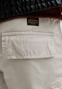 Poche de pantalon beige clair avec des coutures visibles et une étiquette de marque Jack & Jones noire au-dessus de la poche, porté avec une veste noire et marron.