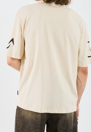 Osoba ubrana w luźny beżowy t-shirt z czarnymi graficznymi elementami na rękawach oraz wyblakłe czarne szorty jeansowe, stoi tyłem do kamery.