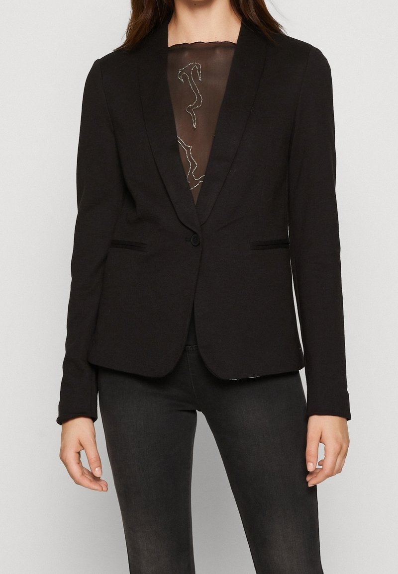 Svart figursydd blazer med slag, enkel knap stängning och två framfickor, över ett genomskinligt, mönstrat plagg.