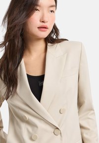 Blazer beige doppiopetto con finitura testurizzata, caratterizzato da due bottoni rotondi e una tasca sul petto. Indossato sopra una maglietta nera.