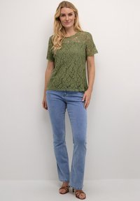 Top in pizzo verde oliva con maniche corte e orlo a scallop, indossato sopra jeans svasati blu chiaro. Il tessuto presenta intricati motivi floreali.