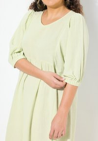 Ljust grön klänning i texturerad tyg. Har en v-ringning, puffärmar med elastiska manschetter och en rynkad midja för en avslappnad passform.