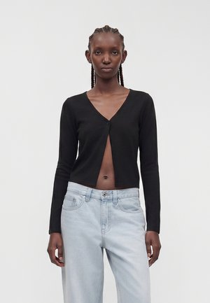 Femme aux cheveux tressés porte un cardigan noir court boutonné une fois au niveau de la poitrine, associé à un jean taille haute bleu clair, fond neutre.