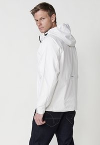 Witte lichtgewicht hooded jas met een gladde textuur, zwarte ritsaccenten en verstelbare trekkoordjes. Gepaard met donkere denimspijkerbroeken.