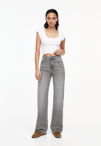 Jeans grii evazați cu talie medie și un top alb scurt. Jeansii au o textură estompată și un sistem de închidere cu buton. Încălțămintea maro completează ținuta.