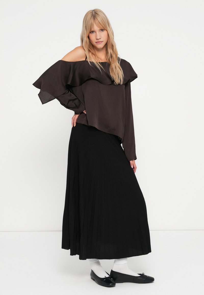 Haut marron à épaules dénudées avec un col volanté, associé à une longue jupe plissée noire. Des ballerines noires et des chaussettes blanches complètent la tenue.