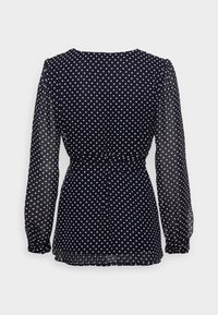 Tmavě modrá dlouhá blůza s bílými puntíky, průhlednými rukávy a stáhnutým pasem, zobrazená zezadu na jednobarevném pozadí.