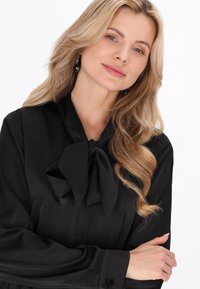 Blusa negra de manga larga con un gran detalle de lazo en el cuello. Tejido liso, puños con botones y un corte holgado.