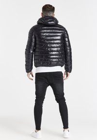 Sort dunjakke med vandret quiltet mønster, skinnende tekstur og hætte. Båret med sorte jeans og hvide sneakers.