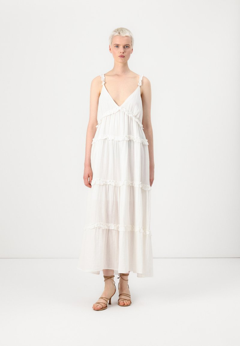 Hvit, lagdelt maxi-kjole laget av lett stoff, med dyp V-hals og blomstertrim på stroppene. Kombinert med sandfargede stroppehæler.