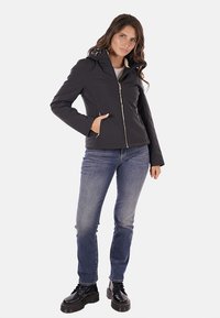 Giacca nera con zip e cappuccio, con una superficie texture e tasche laterali. Abbinata a jeans blu e stivali neri con lacci.