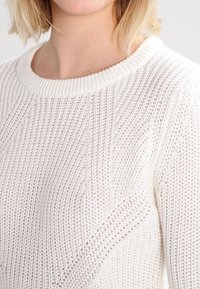 Eine Nahaufnahme einer Frau, die einen weißen, strukturierten Pullover mit weitem Halsausschnitt trägt, wobei der Fokus auf dem Strickmuster des Stoffes und ihrer Schulter liegt.