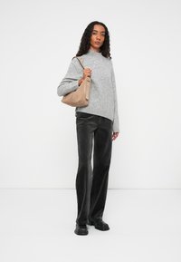 Maglione grigio a maglia con collo alto, abbinato a pantaloni neri in corduroy a zampa d'elefante. Tiene una borsa a tracolla in camoscio color nocciola, in piedi su uno sfondo bianco.