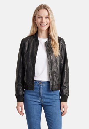 Woman smiling wearing a black leather jacket over a white shirt and blue jeans, standing against a plain light background.