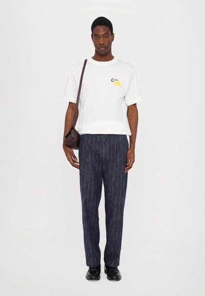 Witte katoenen t-shirt met zwart en geel graphic, gecombineerd met blauw gestreepte broek en zwarte schoenen. Model houdt een kleine bruine crossbody tas vast.