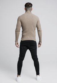 Beige ribbed Rollkragenpullover, kombiniert mit schwarzen Slim-Fit-Jeans und weißen Sneakern, von hinten auf einem neutralen Hintergrund betrachtet.