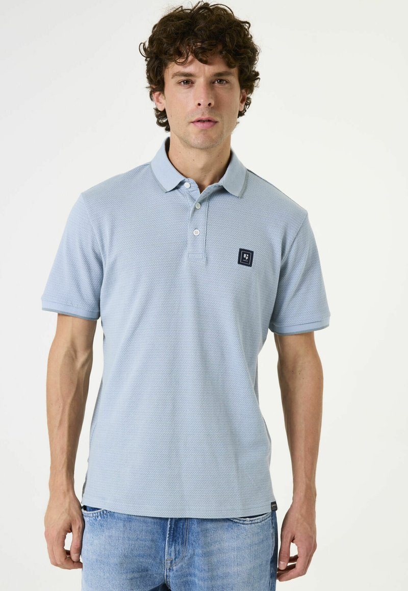 Camisa polo azul claro hecha de tejido texturizado, con cuello, dos botones y un pequeño parche con logo oscuro en el pecho.