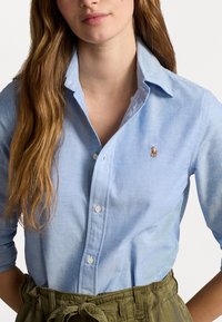 Ljusblå skjorta med knapp-up av tyg med texturerad yta, med en liten broderad logotyp ovanför vänster bröst.