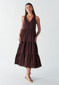 Brun linned maxikjole med V-hals, knapper foran og lagdelt nederdel. Har tynde stropper og en flydende kant. Bæres med hvide sandaler.