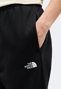 Main dans la poche d'un pantalon de survêtement noir avec taille élastique et logo blanc The North Face sur la cuisse, sur fond uni.