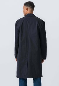 Mand, der vender væk, iført en lang sort frakke og blå jeans mod en ensfarvet lys grå baggrund.