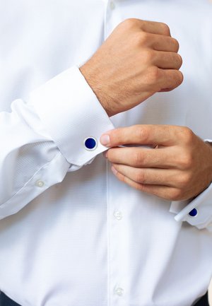 Weißes Hemd mit strukturiertem Gewebe und blauen Manschettenknöpfen in kreisförmigen Fassungen. Hände, die die Manschette anpassen.