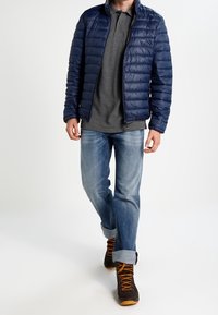 Marinblå quiltad jacka, grå polo-tröja, ljusblå jeans och bruna vandringsskor med orange detaljer. Fokusera på lager-på-lager och utomhusstil.