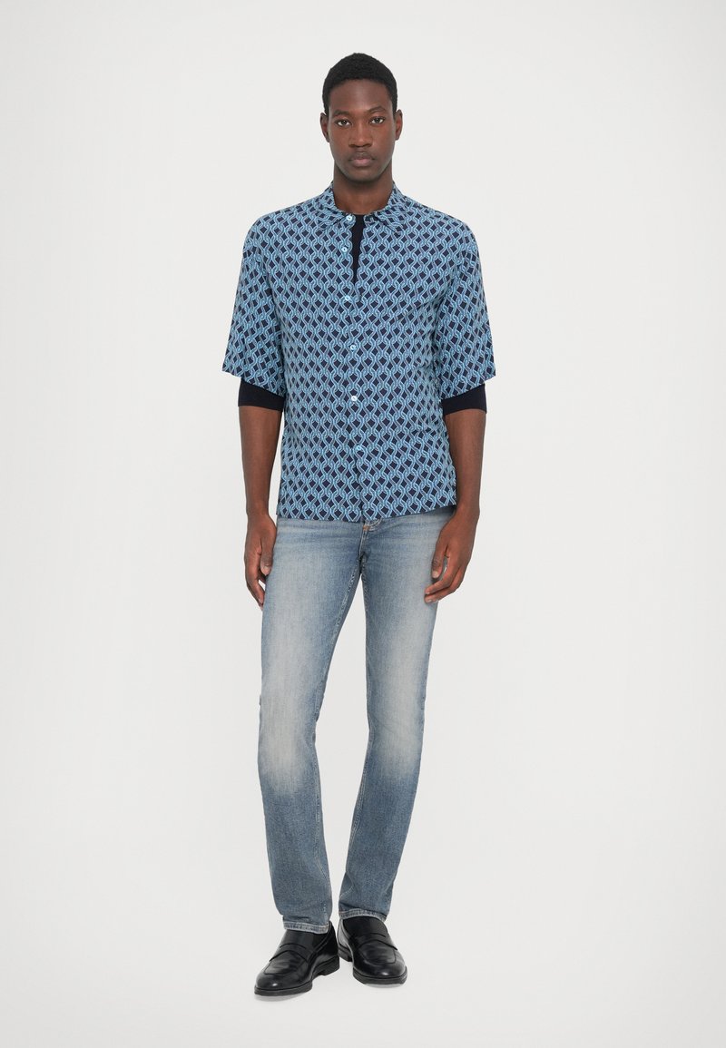 Hombre de pie vistiendo una camisa de manga corta azul con patrón sobre una camiseta de manga larga negra, jeans de color azul claro y zapatos de cuero negros, sobre un fondo blanco.